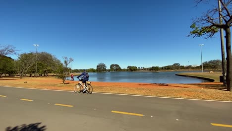 Zeitlupenaufnahmen-Eines-Radfahrers,-Der-Entlang-Eines-Großen-Sees-Im-Stadtpark-Von-Brasilia-Fährt-Und-Einen-Sommertag-Genießt