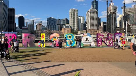 Wide-shot-capturing-tourists-visiting-the-iconic-BRISBANE-block-letter-monumental-sign-at-downtown-southbank-neighborhood-with-high-rise-buildings-and-office-towers-in-the-background,-QLD-Australia