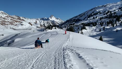 Gente-Divirtiéndose-En-Trineo-En-Un-Campo-De-Nieve-Con-Los-Alpes-Al-Fondo