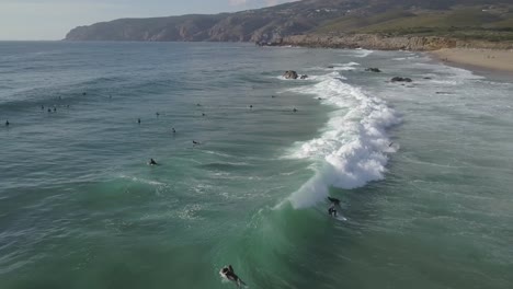 Aerial,-pan-drone-shot-following-surfers-on-the-waves-of-the-atlantic-ocean,-on-a-partly-sunny-evening,-at-the-coast-of-Cascais,-in-Portugal