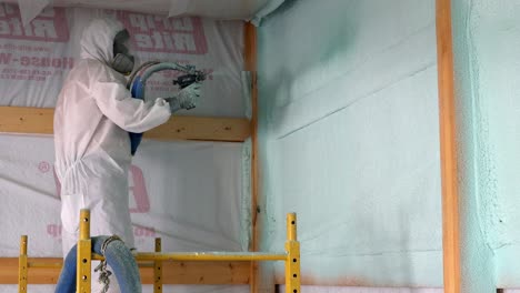 Worker-in-respirator-and-Tyvek-suit-sprays-second-layer-of-closed-cell-foam-insulation-on-the-house-wrap-of-newly-constructed-exterior-wall