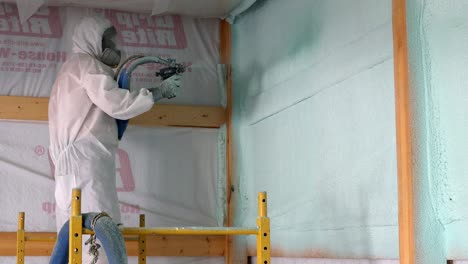 Trabajador-Con-Respirador-Y-Traje-Tyvek-Rocía-Una-Segunda-Capa-De-Aislamiento-De-Espuma-De-Celda-Cerrada-En-La-Envoltura-De-La-Casa-De-Una-Pared-Exterior-Recién-Construida