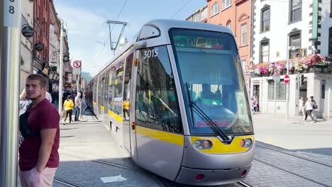 The-Dublin-tram-arrives-at-the-Abbey-Street-stop,-with-many-people-walking-through-the-streets-on-a-sunny-day