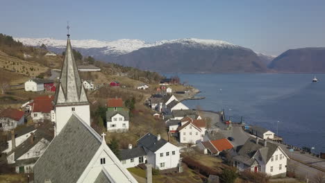 Luftaufnahme-Umkreist-Einen-Kirchturm-In-Einem-Norwegischen-Dorf-Mit-Schneebedeckten-Bergen-Und-Einer-Fähre,-Die-Im-Hintergrund-Einen-Fjord-überquert