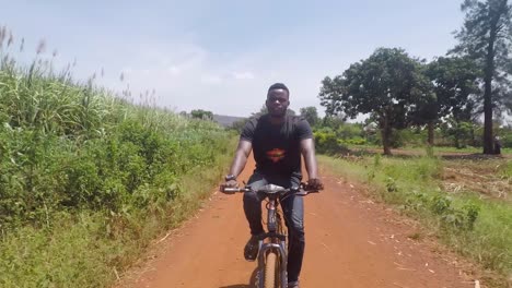 Una-Toma-Estabilizada-De-Un-Hombre-Africano-Montando-Su-Bicicleta-A-Través-De-Aldeas-Rurales