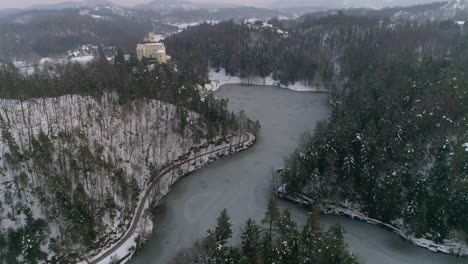 Revealing-shot-of-frozen-lake-surrounded-with-forest-and-fairy-tale-castle-in-the-distance-at-winter