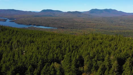 Aerial-drone-shot-over-lush-green-forest-revealing-vibrant-autumn-colors-over-the-ridge-and-a-still-forest-lake-in-the-Maine-wilderness