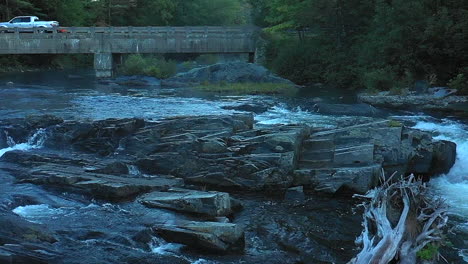Aerial-drone-shot-in-slow-motion-emerging-from-a-thick-mist-over-the-dark-waterfalls-at-Big-Wilson-Falls-and-a-bridge-with-cars-and-trucks-in-the-Maine-wilderness