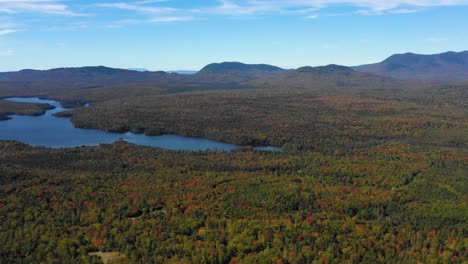 Aerial-drone-shot-panning-right-over-a-large-mountain-lake-in-the-forest-revealing-a-long-mountain-range-as-summer-ends-and-the-season-changes-to-fall-in-Maine