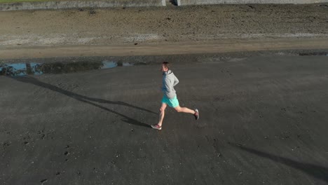 Aerial-tracking-shot-of-young-man-jogging-on-a-beach-in-Auckland,-New-Zealand