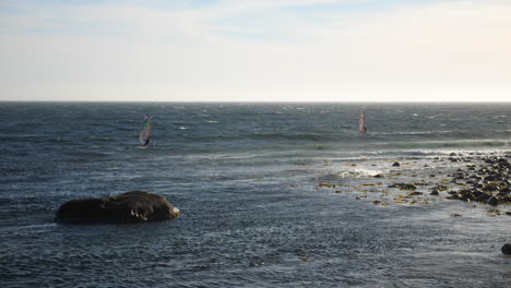 Static-wide-shot-of-norwegian-windsurfers,-surfing-a-waves,-at-the-coast-of-the-North-sea,-in-the-atlantic-ocean,-on-a-sunny,-summer-day,-in-Lista,-South-Norway