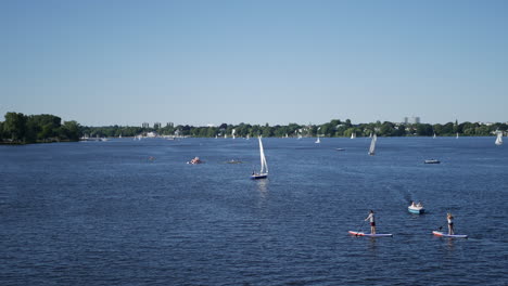 People-enjoy-the-sun-with-stand-up-paddling-and-driving-rowing-boats,-sailboats-and-pedal-boots-on-Outeralster-Lake-in-Hamburg,-Germany-at-fine-weather