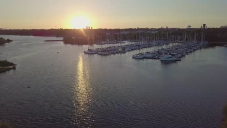 Aerial-Sunset-Rising-Shot-Of-Sailboat-Marina-Yacht-Club-Dock-In-Lake-Bay-Among-Green-Trees-And-Close-Birds-With-City-Buildings-Skyline-In-Background-In-Toronto-Ontario-Canada