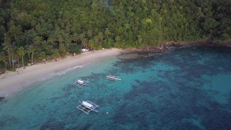 Aerial-view-of-filipino-boats-on-turquoise-waters-and-beautiful-sandy-beach-with-palm-trees-in-the-Philippines---camera-tracking-pedestal-down