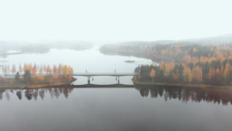Aerial,-drone-shot,-flying-towards-a-bridge,-surrounded-by-mist-and-autumn-colors,,-on-a-foggy,-misty-and-cloudy,-fall-day,-at-lake-Pielinen,-in-Nurmes,-North-Karelia,-Finland