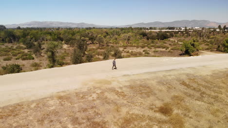 Swooping-drone-and-portrait-shot-of-man-walking-along-path-on-ridge-overlooking-forrest-and-mountains