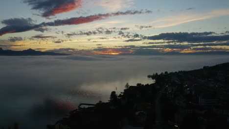 Aerial-passing-over-lake-shore-and-descending-towards-fog-patches-reflecting-in-the-water-at-sunset-Cully,-Lavaux---Switzerland