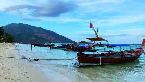 Ein-Filmmaterial-Von-Einem-Strand-Mit-Thailändischen-Booten,-Weißem-Sand-Im-Vordergrund-Und-Einem-Berg-Im-Hintergrund-Mit-Menschen,-Die-Vom-Boot-Aussteigen