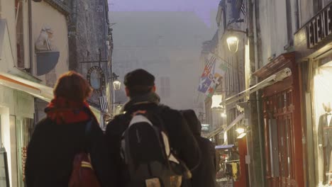 Couples-walk-down-a-warmly-lit-cobblestone-shopping-street-in-an-old-small-town-in-the-Pyrenees-mountains-on-a-foggy-night-in-slow-motion