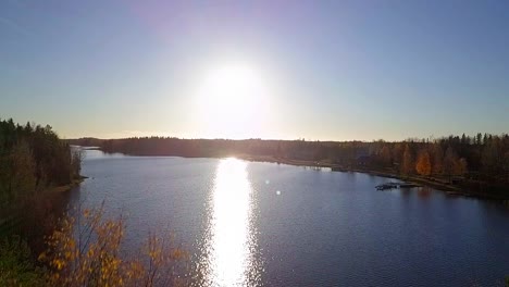 Aerial-ascending-close-to-trees-revealing-idyllic-rural-lake-village-lit-up-by-autumn-sunset,-Ostrobothnia,-Finland