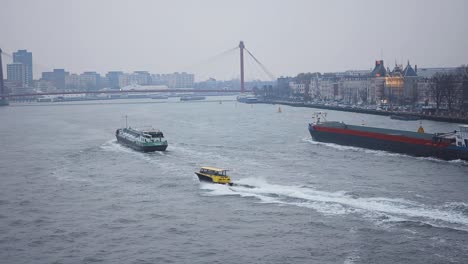 Water-taxi-speeding-on-the-river-Maas-in-between-cargo-ships-with-a-bridge-and-the-skyline-of-the-historical-port-area-of-Rotterdam-in-the-background-on-a-drowsy-overcast-day