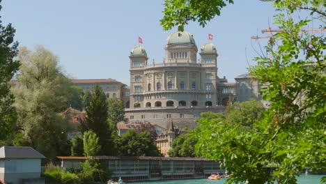 waving-swiss-flags-on-the-rooftop-of-the-Bundeshaus-in-Bern,-Switzerland-from-the-Aare-river-site-on-a-windy-day-ZOOM-IN-wide-low-angle-shot
