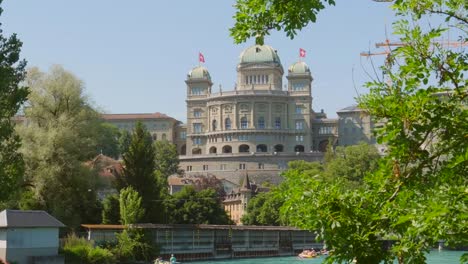 Ondeando-Banderas-Suizas-En-La-Azotea-Del-Bundeshaus-En-Berna,-Suiza-Desde-El-Sitio-Del-Río-Aare-En-Un-Día-Ventoso-Zoom-En-Tiro-De-ángulo-Bajo-Amplio