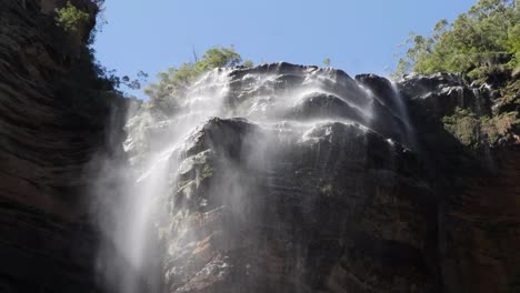 Low-angle-view-of-wentworth-falls-in-the-blue-mountains,-Australia-with-the-wind-blowing-the-water-coming-down-and-blue-sky