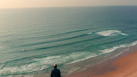 Aerial:-A-man-standing-on-a-viewpoint-watching-the-surfers-in-Portugal