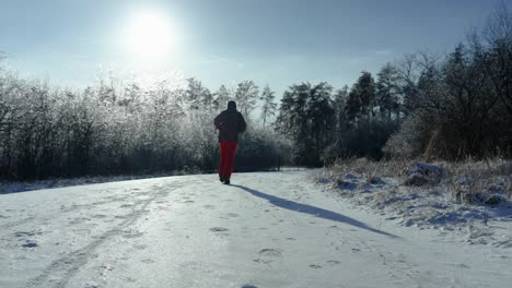Ein-Mann-Läuft-An-Einem-Schönen-Sonnigen-Und-Kalten-Winternachmittag-Auf-Einem-Schneebedeckten-Und-Eisbedeckten-Waldweg-Von-Der-Kamera-Weg