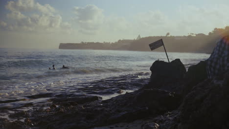 Two-young-surfers-begin-to-paddle-out-in-the-water-during-the-early-morning-in-Bingin-Beach-Bali-as-a-flag-blows-in-the-foreground