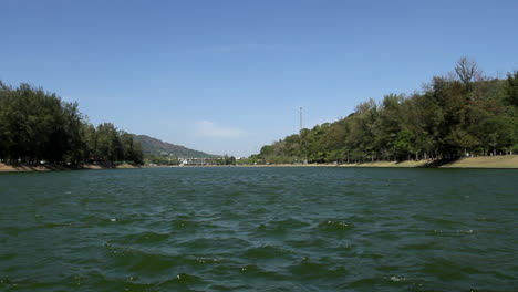 locked-low-angle-shot-of-panoramic-view-of-Nai-Harn-Lake-on-a-breezy-blue-sky-summer-day,-no-people,-water-in-foreground-and-idyllic-park-and-forest-in-background