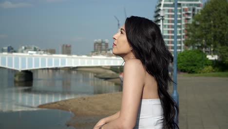 Beautiful-latina-woman-on-holiday-leaning-against-the-railing,-looking-at-the-river-Thames-in-London-pointing-up-at-the-sky,-smiling-and-wandering