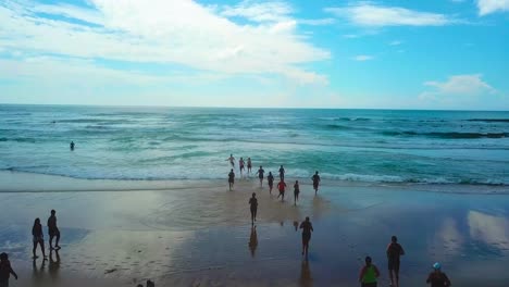 Aerial-tracking-view-of-beach-and-people-running-into-a-blue-ocean