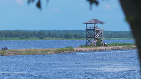 View-of-people-walking-on-footbridge-path-and-birdwatching-tower-at-lake-Liepaja-in-sunny-summer-day-with-scenic-cumulus-clouds,-medium-shot-from-a-distance
