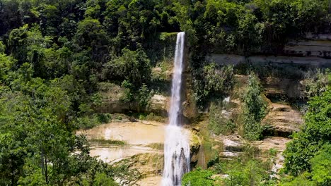 A-beautiful-waterfall-cascades-down-a-cliff-into-a-beautiful-turquoise-pool-below-in-the-middle-of-a-lush-green-jungle-in-Bohol,-Philippines-while-locals-enjoy-the-refreshing-cool-water