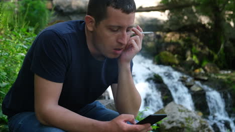 Young-man-holding-and-smoking-a-cigarette-while-browses-social-media-on-a-smartphone-near-a-waterfall