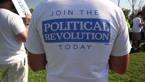 Slow-dolly-zoom-in-shot-of-back-of-Caucasian-male-T-shirt,-zoom-to-closeup-of-"Join-the-Political-Revolution-today"-as-people-gather-for-political-rally-in-San-Jose,-CA