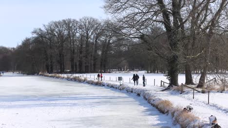 People-walking-along-and-ice-skating-on-the-frozen-river-Berkel-near-Hanseatic-city-Zutphen-in-winter-snow-landscape-with-winter-barren-trees-in-the-background