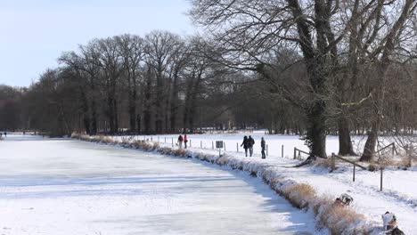 Menschen,-Die-Entlang-Gehen-Und-Eislaufen-Auf-Dem-Zugefrorenen-Fluss-Berkel-Bei-Der-Hansestadt-Zutphen-In-Winterlicher-Schneelandschaft-Mit-Winterkargen-Bäumen-Im-Hintergrund