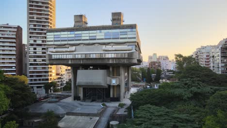 Aerial-pan-right-of-brutalist-style-National-Library-between-trees-and-buildings-at-sunset,-Buenos-Aires