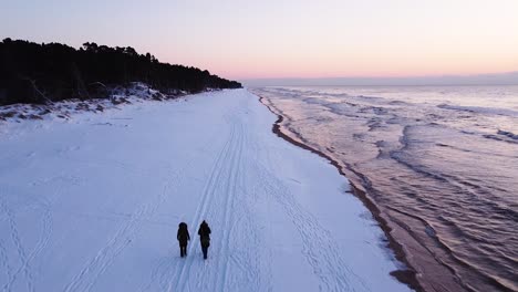 Beautiful-romantic-aerial-shot-flying-over-a-snow-covered-beach-and-couple-after-the-sunset-on-the-calm-Baltic-sea-coastline,-high-contrast-sky,-wide-angle-ascending-drone-shot-moving-forward-fast