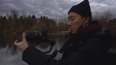Man-using-a-DLSR-camera-mounted-on-a-gimbal-to-record-film-scenery-of-a-lake-at-a-park-with-trees-clouds-sky-reflecting-off-of-water-in-the-background