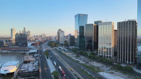 Aerial-dolly-in-of-boats-in-Puerto-Madero-docks-near-Paseo-del-Bajo-highway-and-skyscrapers-at-sunset,-Buenos-Aires