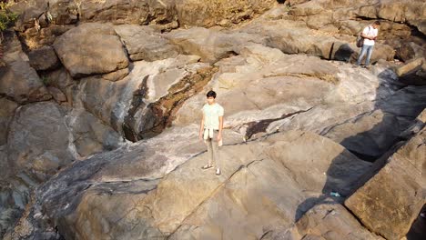 A-Drone-view-of-a-young-Indan-boy-standing-on-edge-of-a-small-hill-closeup-to-landscape-shot-video-background,-Aerial-view-of-a-small-hill-and-dried-waterfall-with-rocks-and-stones-in-India