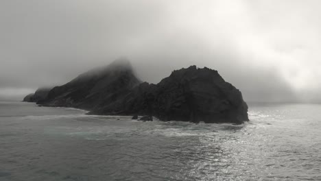 Big-rock-shrouded-in-mist-in-open-sea-near-Ponta-da-Calheta-beach,-Portugal