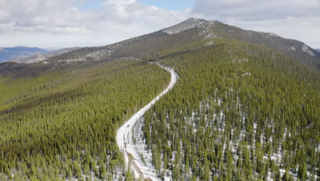 Aerial-reveal-forward-of-sunny-and-snow-covered-back-roads-in-the-Colorado-mountains-surrounded-by-bright-green-pine-tree-forests-with-blue-skies