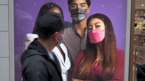 Fußgänger,-Die-Schützende-Gesichtsmasken-Als-Vorbeugung-Gegen-Das-Covid-19-virus-Tragen,-Gehen-Auf-Der-Straße-An-Einer-Kommerziellen-Werbung-Einer-Marke-Für-Gesichtsmasken-In-Hongkong-Vorbei