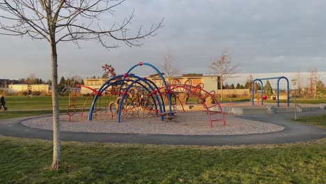 Rotating-around-a-colourful-metallic-playground-kid-play-are-in-local-community-park-grass-field-and-people-walking-in-the-background