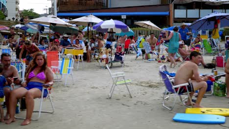 Dolly-left-of-families-sitting-under-umbrellas-in-the-sand-near-the-shore-of-Bombas-and-Bombinhas,-Brazil